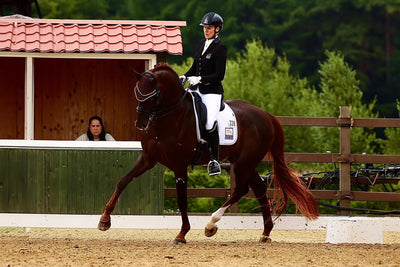 Bernadette Brune trotting her horse in a dressage test at an outdoor show, wearing a JuulC competition jacket